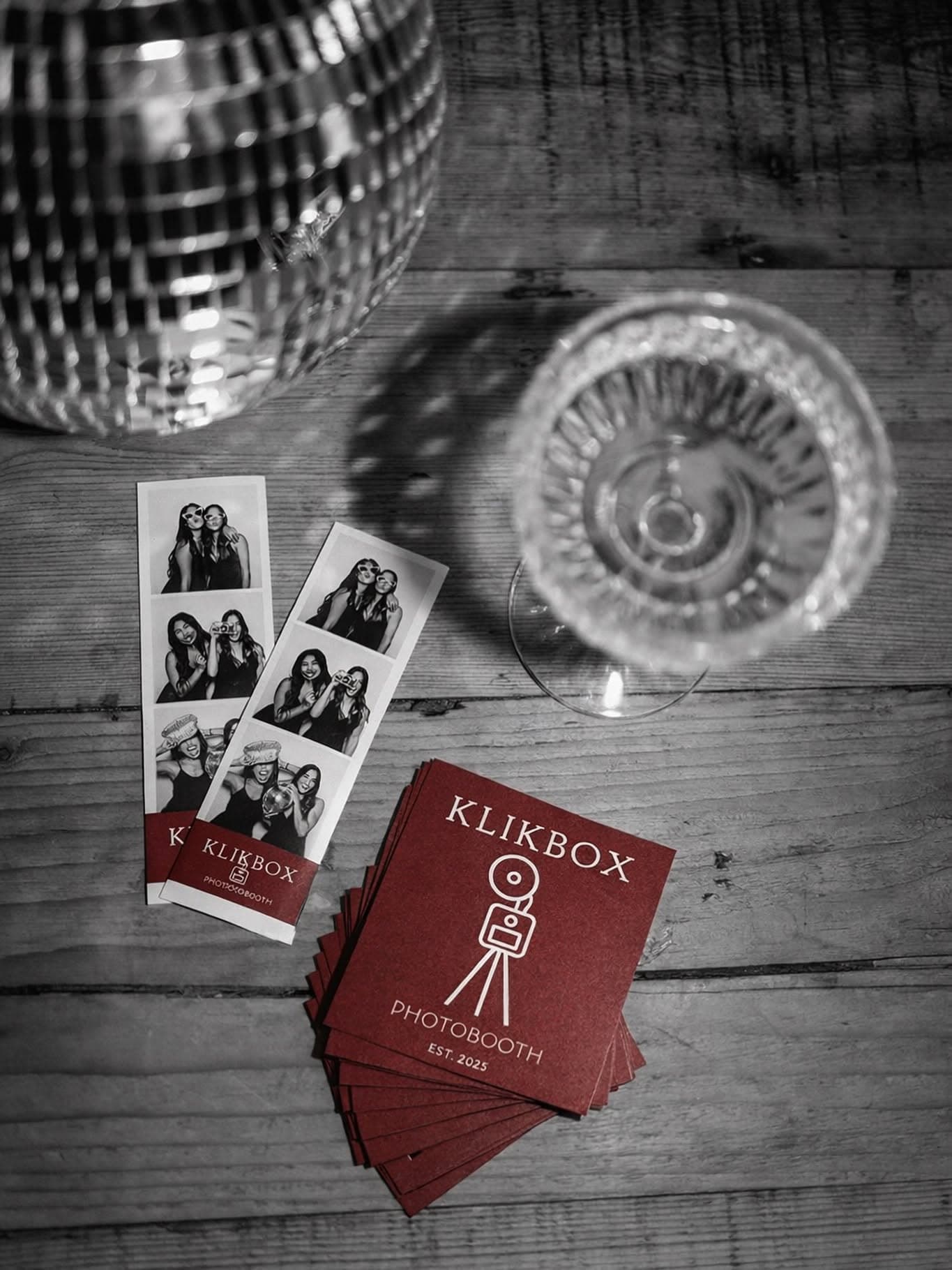 Ασπρόμαυρο flat-lay με εκτυπωμένα photostrips του ΚλικBox, disco ball και ποτήρι σαμπάνιας πάνω σε ξύλινο τραπέζι